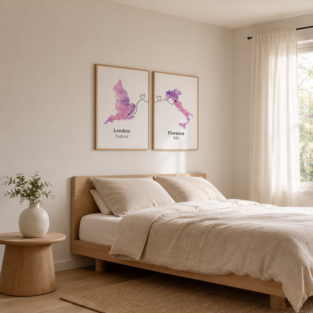 A styled bedroom scene with a light wood bed dressed in cream bedding, a small wooden side table with a white vase of greenery, and two framed art prints above the bed. The prints show watercolor-style maps of London and Florence in pink and purple tones.