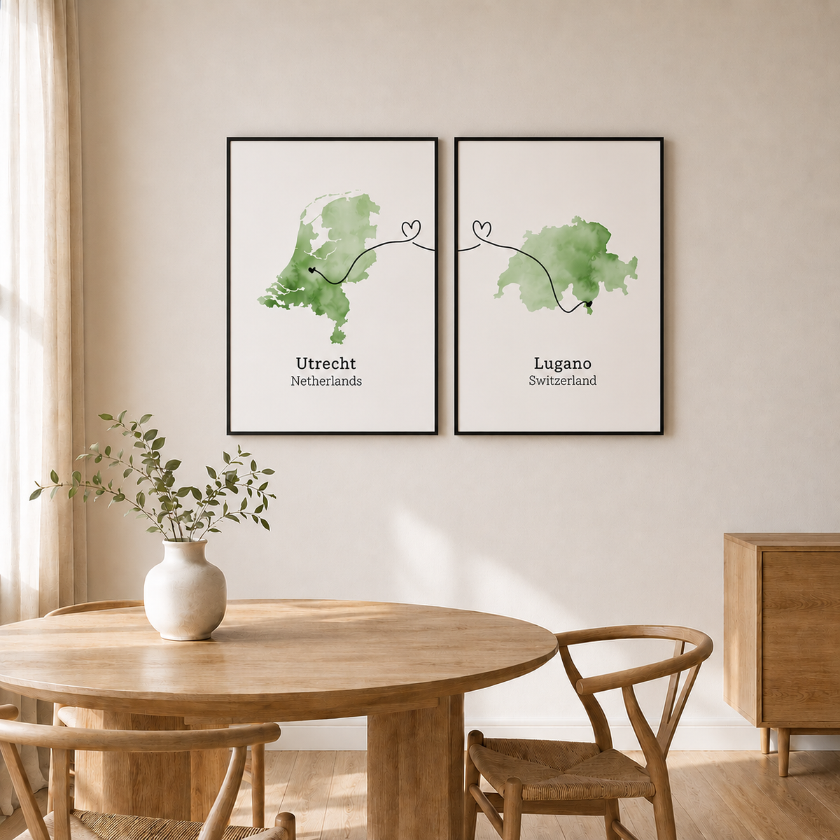 Two framed watercolor map art prints hanging on a beige wall above a round wooden dining table with chairs. The left print shows Utrecht, Netherlands in green, and the right print shows Lugano, Switzerland, also in green.