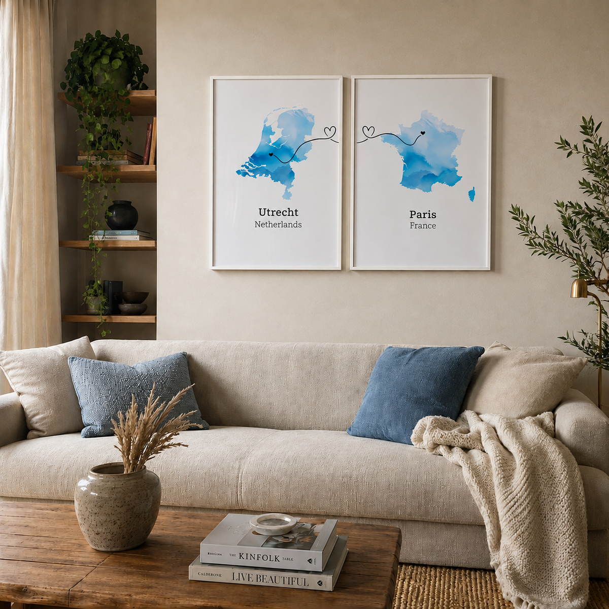 A styled living room scene with a beige sofa, blue throw pillows, a wooden coffee table, and two framed wall art prints hanging above the couch. The prints show blue watercolor maps of Utrecht and Paris with small heart motifs connecting them.